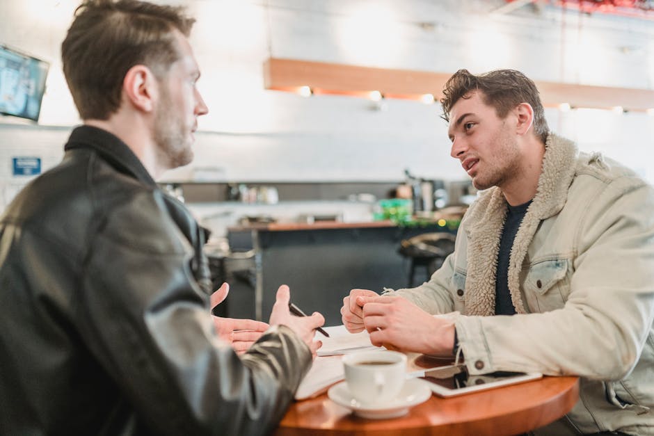 Side view of crop young man talking about business project at table with papers and looking at partner on blurred background